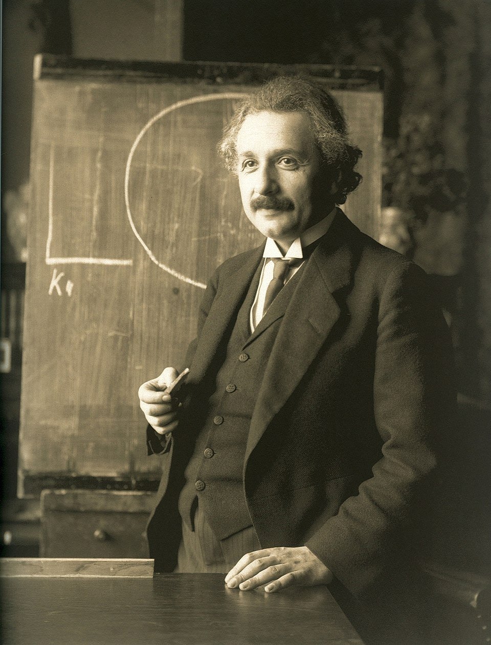 Albert Einstein áll egy táblánál, kezében krétával, tudományos magyarázatot ad.