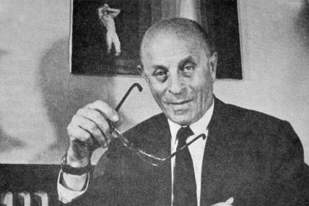 Bíró László József mosolyogva ül, kezében szemüveget tartva, háttérben festmény látható.