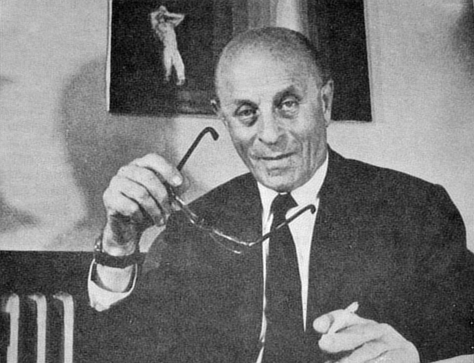 Bíró László József mosolyogva ül, kezében szemüveget tartva, háttérben festmény látható.