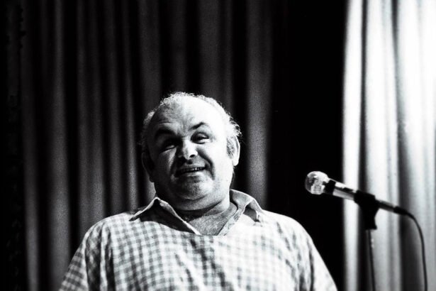 Egy mosolygó férfi áll a mikrofon előtt, háttérben függöny.