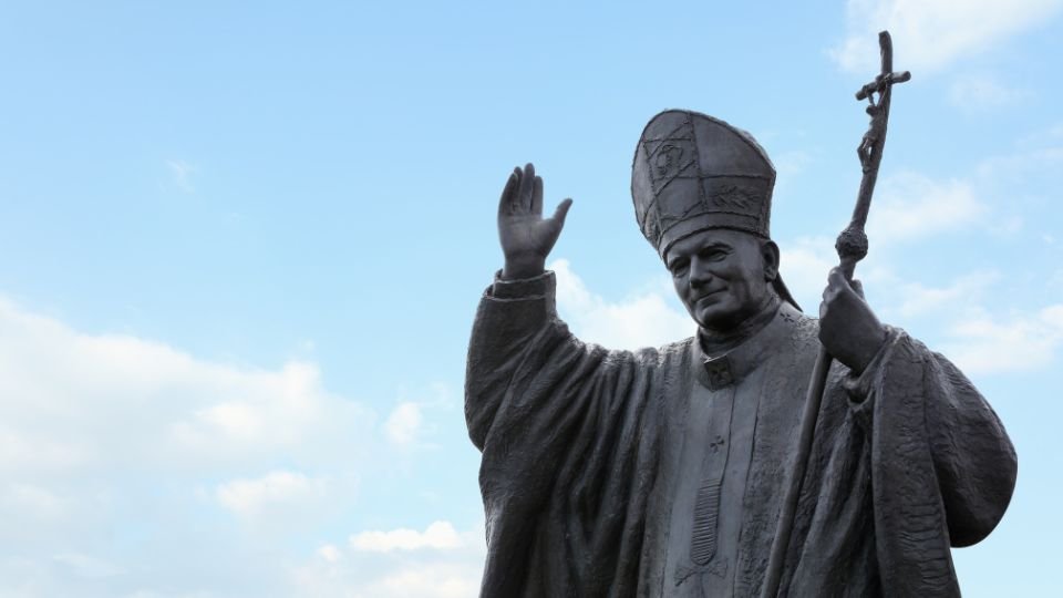 II. János Pál pápa szobra, amely integet a látogatóknak egy világos égen.