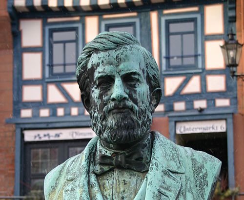 Johann Philipp Reis mellszobra, bronz színű, részletes kidolgozással, háttérben régi épületekkel.