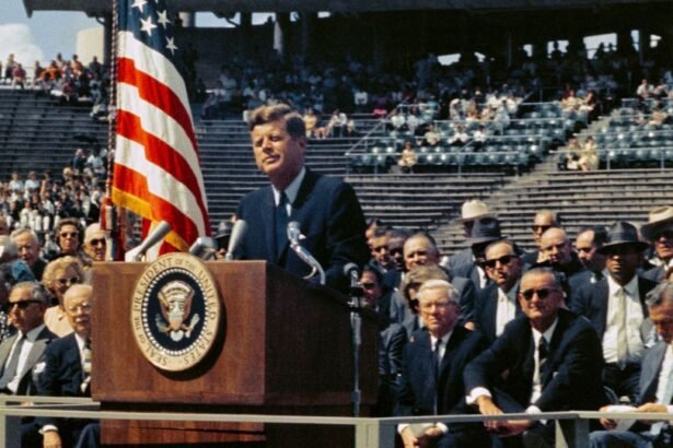 John F. Kennedy elnök beszédet mond egy rendezvényen, háttérben amerikai zászló.