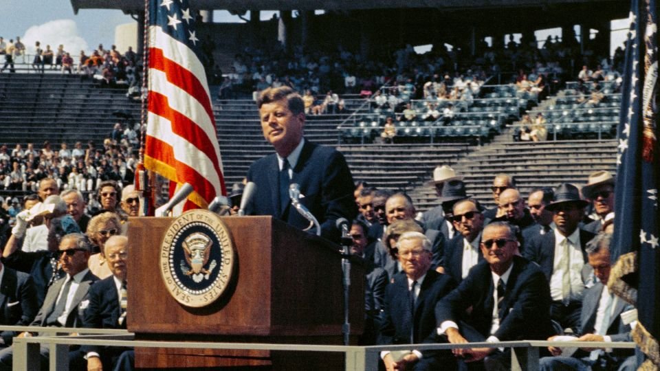 John F. Kennedy elnök beszédet mond egy rendezvényen, háttérben amerikai zászló.
