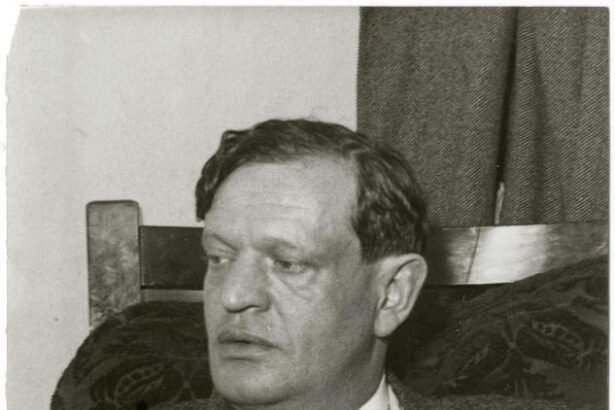 Karinthy Frigyes ül egy karosszékben, komoly arckifejezéssel, a háttérben függöny látható.