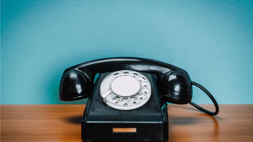 Régi fekete telefon, fehér tárcsával, faasztalon, kék háttér előtt.