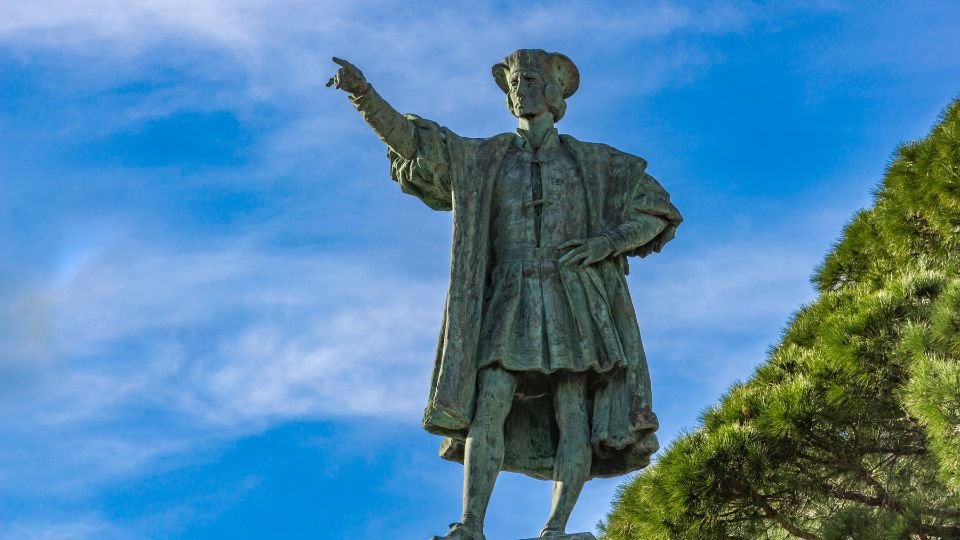 Kolumbusz Kristóf szobra, amely az égre mutat, háttérben kék ég és fák.