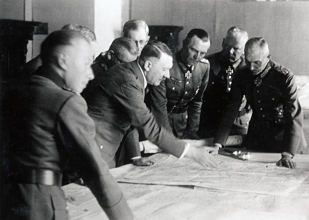 Több férfi egy térképen dolgozik, Adolf Hitler középen mutat.