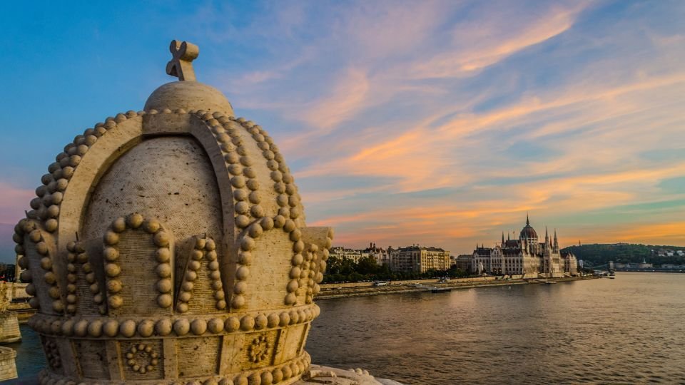 A budapesti Parlament és a Duna látképe naplementében, a koronás díszítéssel előtérben.
