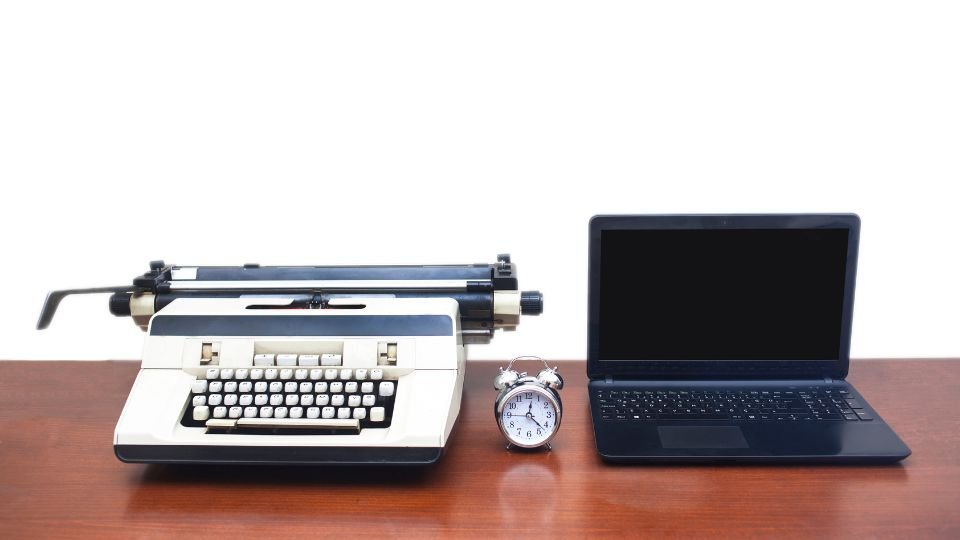 Egy régi írógép, egy laptop és egy óra egy asztalon, a technológiai fejlődés szimbólumai.