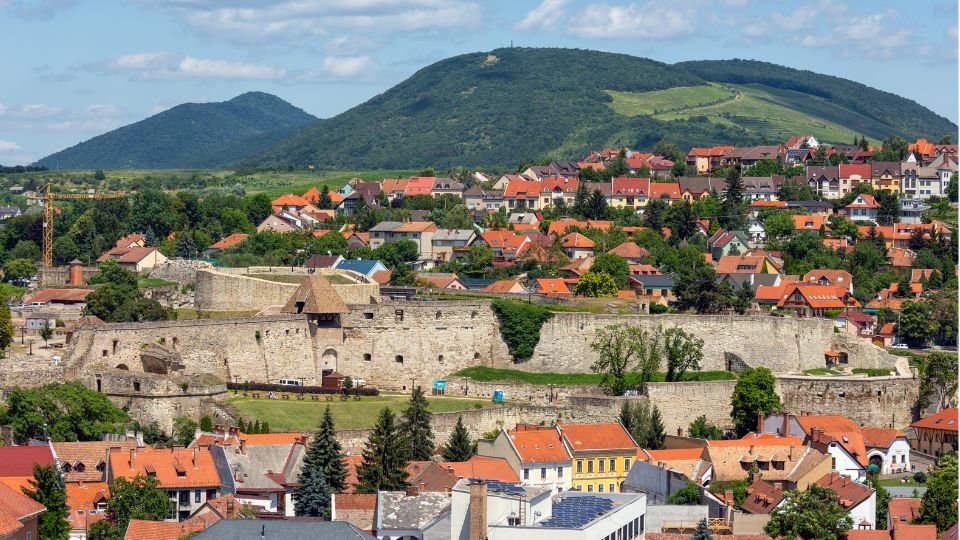 Középkori vár látképe, zöld dombokkal és város házaival körülvéve.