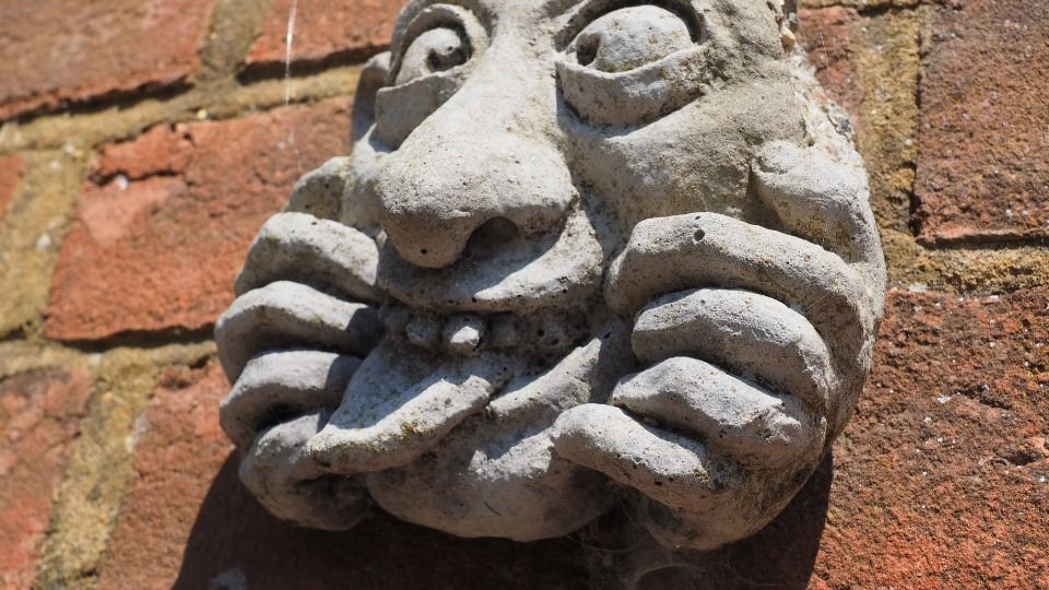 Egy groteszk kőszobor, amely mosolygó arcot formáz, kezével az arcát fogja.