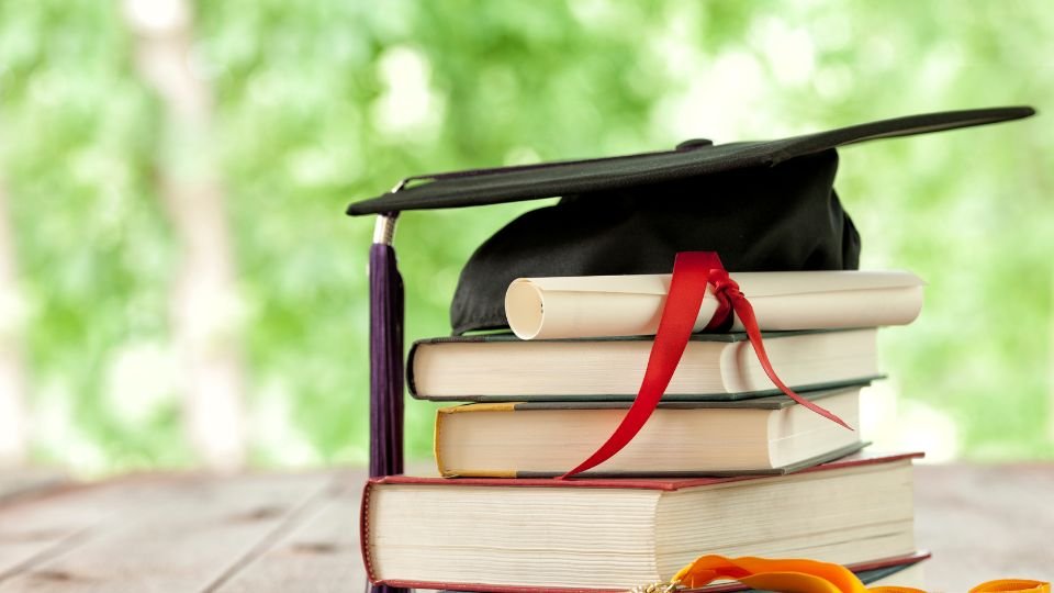 Könyvek, diploma és kalap egy asztalon, zöld háttér előtt.